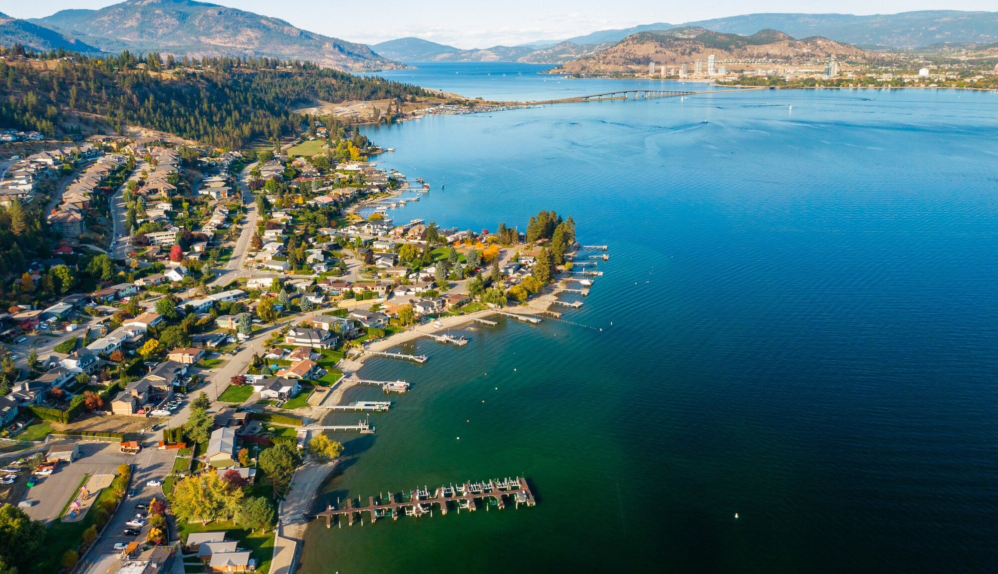 Aerial view over West Kelowna homes and docks on Okanagan Lake, looking toward Kelowna and the Interior mountains