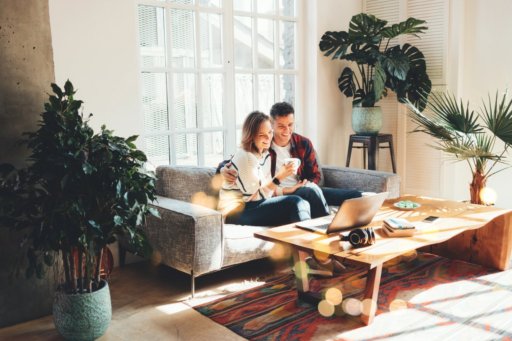 A bright, plant-filled apartment living room in full sunlight, a couple relaxing on the sofa with coffee and a laptop