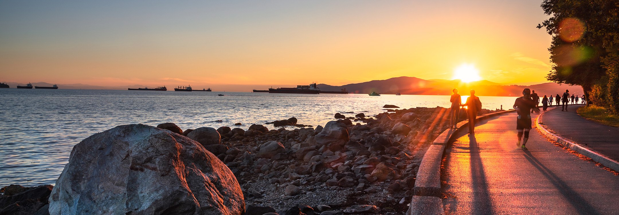 People walking and jogging the Stanley Park seawall at sunset, freighters at anchor in English Bay